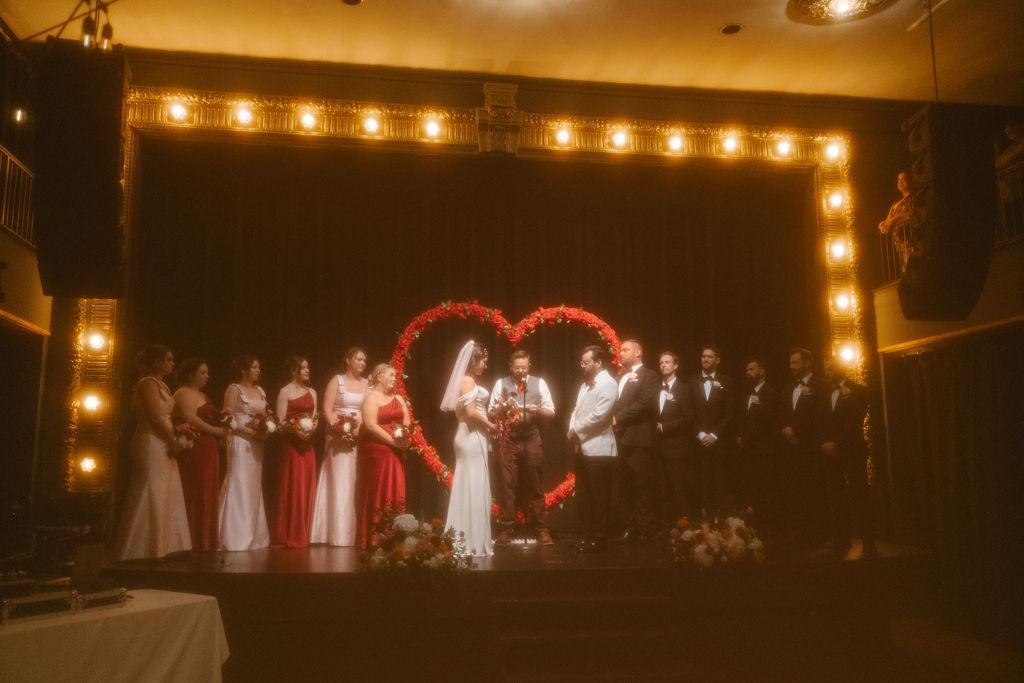 Couple standing on the woodwards stage with their bridal party during the ceremony, with a large heart shaped backdrop behind them. Photo by Photo By Hannah Stump Photography.